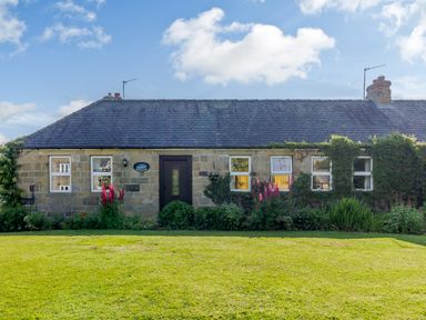 A stone cottage with a dark roof and a garden with flowers and greenery at Barnacre in Longhirst near Morpeth