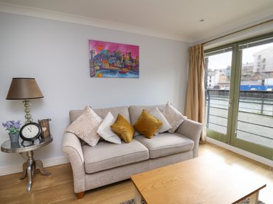 A living room with a beige sofa with cushions a wooden coffee table a side table with a lamp clock and flowers and large glass doors at Flat 2 in Pembroke