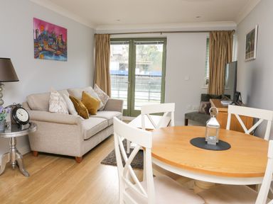 A living room with a beige sofa and cushions a round wooden table with white chairs a television on a wooden stand and double doors with curtains at Flat 2 in Pembroke