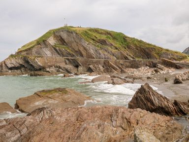 A rocky coastline with waves hitting the shore and a green hill in the background at 65 Channel View in Ilfracombe