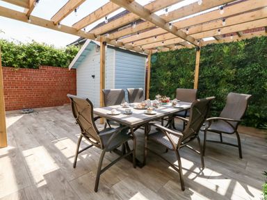 An outdoor patio with a wooden pergola string lights a dining table set with six chairs and tea sets at The Chocolate Box in Weymouth