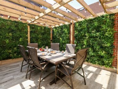 An outdoor dining area with a wooden table and six chairs under a wooden pergola with string lights and green foliage walls at The Chocolate Box in Weymouth