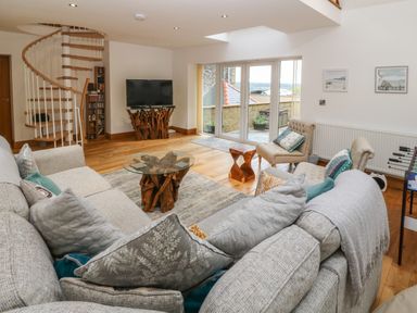 A living room with a spiral staircase a glass top table with wooden base a television two armchairs and a large sofa at On the Straits in Glyngarth near Menai Bridge