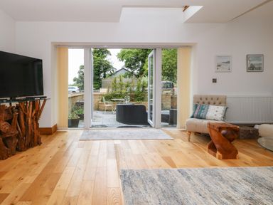 A living room with a wooden TV stand a beige armchair and a small wooden table next to a glass door leading to an outdoor patio at On the Straits in Glyngarth near Menai Bridge