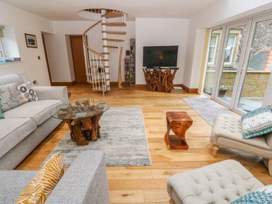 A living room with gray sofas a glass coffee table a wooden side table a spiral staircase a television on a wooden stand and large windows at On the Straits in Glyngarth near Menai Bridge