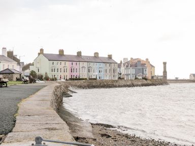 A waterfront promenade with benches and pastel-colored houses along a stone sea wall at Gwel Y Mor in Beaumaris