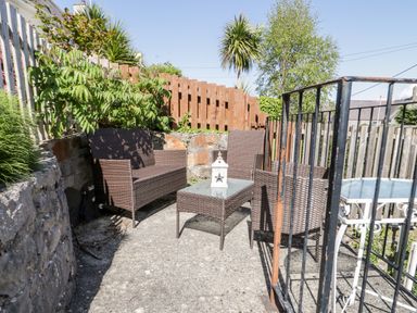 An outdoor seating area with wicker chairs and a glass coffee table next to a wooden fence and stone wall at Gwel Y Mor in Beaumaris