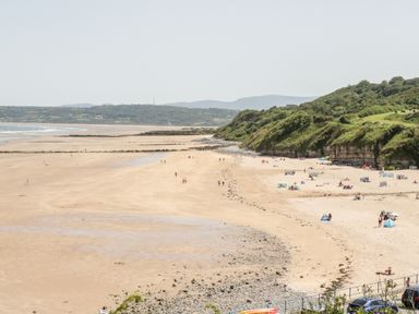 A sandy beach with people walking and sitting near grassy cliffs and hills in the background