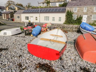 A small red and white boat on a pebble beach with other boats and fishing equipment near houses with solar panels on the roof at Gwel Y Mor in Beaumaris