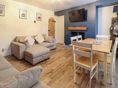 A living room with beige sofas a wooden dining table with white chairs a wall-mounted tv and a fireplace at Gwel Y Mor in Beaumaris