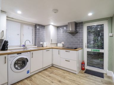 A kitchen with white cabinets wood countertops a washing machine a window with blinds and a door with a fire exit sign at Gwel Y Mor in Beaumaris
