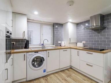 A kitchen with white cabinets, wooden countertops, a washing machine, a sink, a stove, a kettle, a toaster, a knife block, and gray tiled backsplash at Gwel Y Mor in Beaumaris