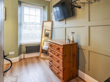 A room with a wooden chest of drawers a full-length mirror leaning against a window with blinds a wall-mounted TV hangers on the wall and a radiator below the window at Gwel Y Mor in Beaumaris