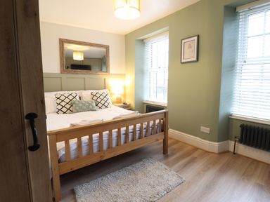 A bedroom with a wooden bed frame white bedding patterned cushions a bedside table with a lamp a wall mirror and two windows with blinds at Gwel Y Mor in Beaumaris