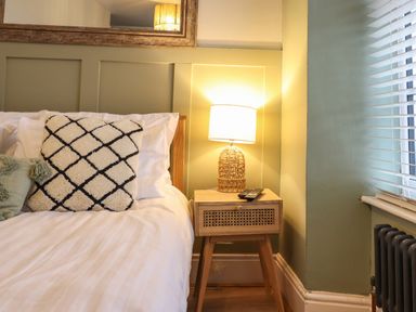 A bedroom corner with a wooden bed with white bedding and patterned cushions next to a wooden nightstand with a lamp and remote at Gwel Y Mor in Beaumaris