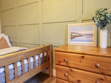 A bedroom with a wooden bed frame, a wooden dresser with a framed picture and a vase with plants on top at Gwel Y Mor in Beaumaris