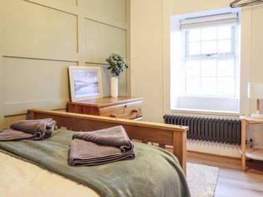 A bedroom with a wooden bed frame, folded towels on the bed, a wooden dresser with a framed picture and vase, a window with a radiator below and a small table with a lamp at Gwel Y Mor in Beaumaris