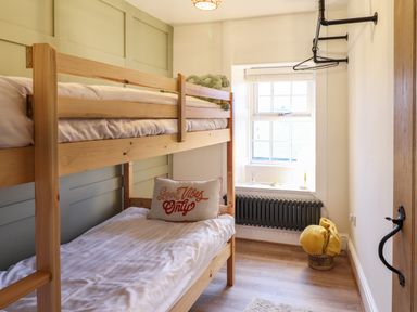 A small bedroom with wooden bunk beds a window a radiator and a basket with yellow cushions at Gwel Y Mor in Beaumaris