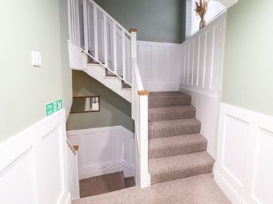 A carpeted staircase with white wooden railings and wainscoted walls with a green upper wall and a fire exit sign on the wall at Gwel Y Mor in Beaumaris