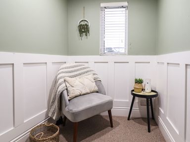 A corner room with a gray chair with a pillow and throw blanket a small black side table with decorative items a wicker basket on the floor and a small window above Gwel Y Mor in Beaumaris