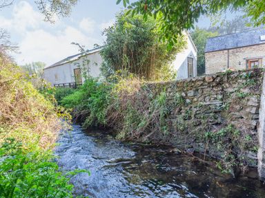 A stream flowing beside a stone wall with plants and two buildings in the background at Millers Lodge in St Keyne near Liskeard