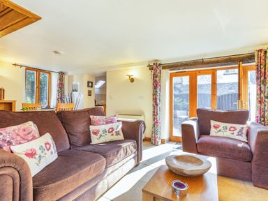 A living room with two brown sofas with floral cushions a wooden coffee table and large windows with floral curtains at Miller's Lodge in St Keyne near Liskeard