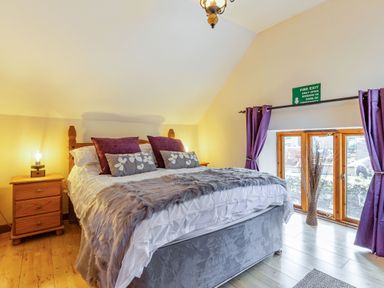 A bedroom with a double bed bedside tables with lamps and a window with purple curtains at Millers Lodge in St Keyne near Liskeard
