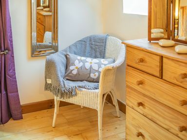 A white wicker chair with a gray throw and floral pillow next to a wooden dresser with a mirror at Millers Lodge in St Keyne near Liskeard