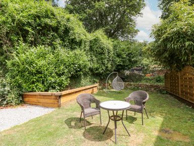A garden with two wicker chairs and a round table on grass surrounded by green bushes and wooden fencing at Millers Lodge in St Keyne near Liskeard