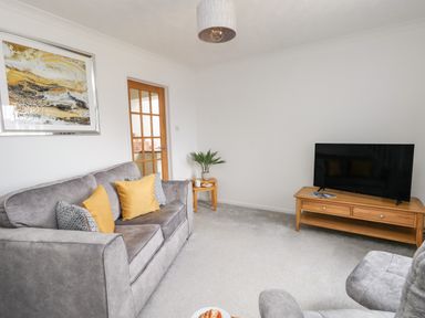 A living room with a gray sofa with cushions a wooden tv stand with a television a side table with a plant and a glass door at Sandpiper in Weymouth