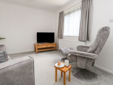A living room with a grey armchair and footstool a wooden side table with a plate and mug a wooden TV stand with a TV and a grey sofa at Sandpiper in Weymouth