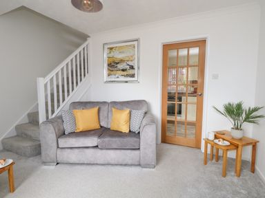 A living room with a grey sofa yellow and patterned cushions wooden tables with a plant and plates stairs and a glass panel wooden door at Sandpiper in Weymouth