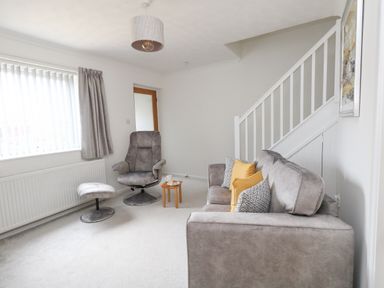 A living room with a grey armchair and footstool near a window and a grey sofa with patterned and yellow cushions near stairs at Sandpiper in Weymouth
