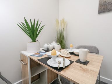 A small dining table set with two egg cups, mugs, plates, and cutlery next to a white cabinet with a potted plant and decorative grass in a corner at Sandpiper in Weymouth