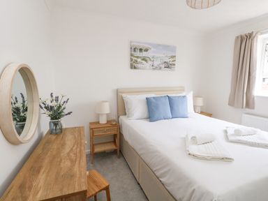 A bedroom with a bed containing blue pillows and rolled towels bedside tables with lamps a wooden desk with a round mirror and flowers at Sandpiper Weymouth