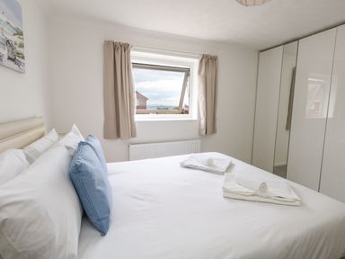 A bedroom with a bed white sheets blue pillows folded towels a window with beige curtains and glossy white wardrobes at Sandpiper in Weymouth