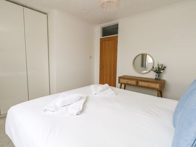 A bedroom with a white bed, folded towels, a wooden door, a white wardrobe, and a wooden table with a round mirror and flowers at Sandpiper in Weymouth
