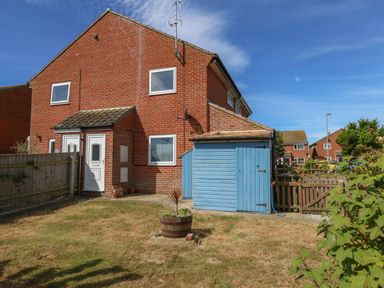 A brick house with white doors and windows a blue wooden shed a wooden fence and a grassy garden with a plant in a planter at Sandpiper in Weymouth