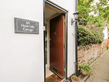 An open wooden front door with a doormat next to a wall sign reading Hock-tide Cottage and a stone wall with plants at Hock-tide Cottage in Kenilworth