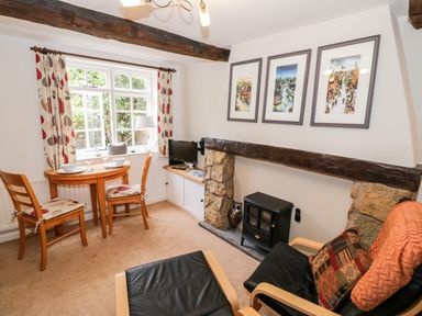 A small dining area with a round wooden table and two chairs by a window and a living area with an armchair and a black stove at Hock-tide Cottage in Kenilworth