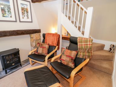 A sitting area with two black leather chairs with wooden frames and patterned cushions near a small stove and carpeted stairs at Hock-tide Cottage in Kenilworth