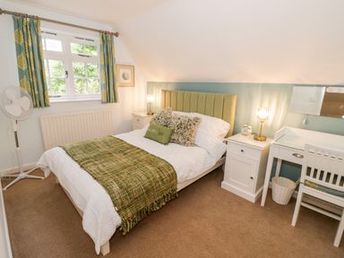 A bedroom with a double bed bedside tables a desk chair window with curtains and a standing fan at Hock-tide Cottage in Kenilworth