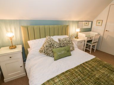 A bedroom with a green upholstered headboard white bedding green cushions bedside tables with lamps and a desk with chair at Hock-tide Cottage in Kenilworth