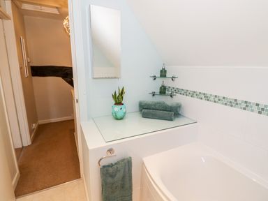 A bathroom corner with a white bathtub green towels a small plant on a glass surface and two glass shelves with soap dispensers at Hock-tide Cottage in Kenilworth