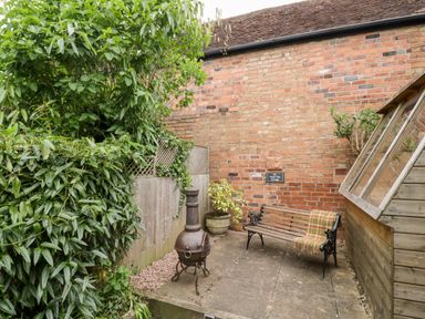 An outdoor patio area with a wooden bench and blanket a metal chiminea a brick wall and plants at Hock-tide Cottage in Kenilworth