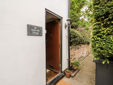 An open wooden door on a white exterior wall with a sign reading 6A Hock-tide Cottage next to potted plants and a stone wall pathway at Hock-tide Cottage in Kenilworth