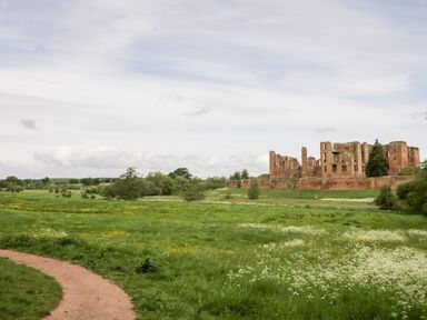 A grassy field with wildflowers a dirt path and the ruins of a large brick building in the background at Hock-tide Cottage in Kenilworth