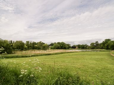 A grassy field with wildflowers near a pond surrounded by trees and houses in the distance at Hock-tide Cottage in Kenilworth
