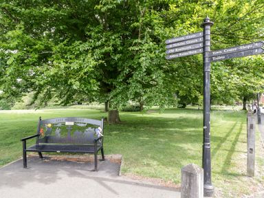 A park area with a memorial bench and a directional signpost near a large tree at Hock-tide Cottage in Kenilworth