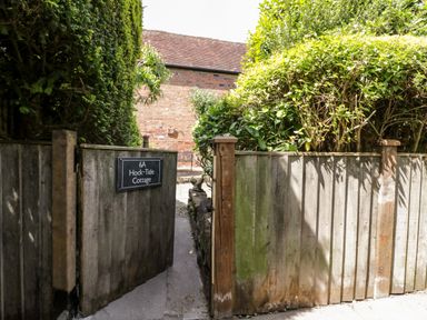 A wooden gate with a sign reading 6A Hock-Tide Cottage surrounded by greenery and a brick building in the background at Hock-tide Cottage in Kenilworth